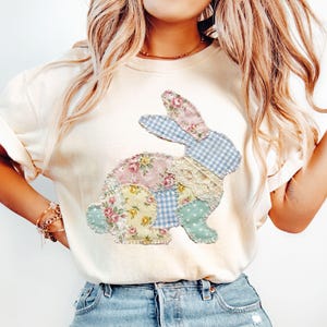 Puede incluir: Camiseta color crema con un diseño de conejo de patchwork. El conejo está compuesto por varios patrones de tela, incluyendo estampados florales, cuadros vichy y lunares. La camiseta tiene mangas cortas y un ajuste relajado, adecuada para uso casual.