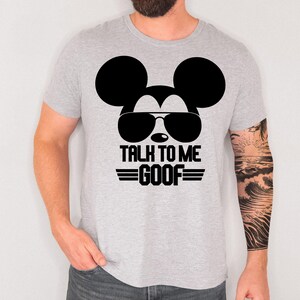 May include: A light gray short-sleeve t-shirt featuring a black graphic of a mouse silhouette wearing aviator sunglasses. Below the graphic, the text reads "TALK TO ME GOOF" in a bold, black, block-style font with horizontal lines on each side.