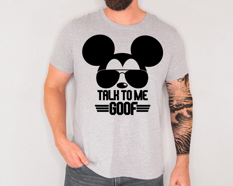 May include: A light gray short-sleeve t-shirt featuring a black graphic of a mouse silhouette wearing aviator sunglasses. Below the graphic, the text reads "TALK TO ME GOOF" in a bold, black, block-style font with horizontal lines on each side.