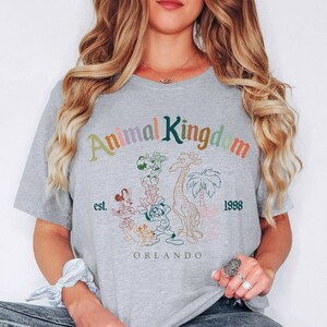 May include: A light gray t-shirt with the words "Animal Kingdom" in colorful text. The shirt features a graphic of cartoon animals, a giraffe, and palm trees. The text "est. 1998 Orlando" is also printed on the shirt.
