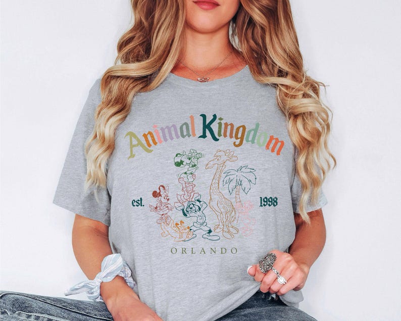 May include: A light gray t-shirt with the words "Animal Kingdom" in colorful text. The shirt features a graphic of cartoon animals, a giraffe, and palm trees. The text "est. 1998 Orlando" is also printed on the shirt.