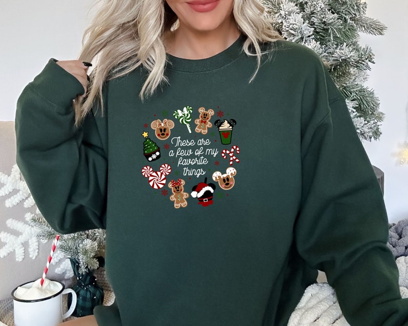 May include: Green Christmas sweatshirt with a festive design featuring Mickey Mouse characters, candy canes, a gingerbread man, a Christmas tree, and a coffee cup. The text on the sweatshirt reads "These are a few of my favorite things."