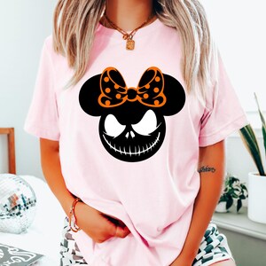 Puede incluir: Una camiseta rosa claro con un gr&aacute;fico negro y naranja. El gr&aacute;fico combina una silueta de Mickey Mouse con una cara de Jack Skellington y un lazo de lunares. La camiseta es de estilo casual y de manga corta.
