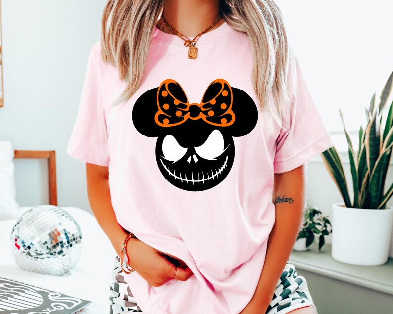 Puede incluir: Una camiseta rosa claro con un gr&aacute;fico negro y naranja. El gr&aacute;fico combina una silueta de Mickey Mouse con una cara de Jack Skellington y un lazo de lunares. La camiseta es de estilo casual y de manga corta.
