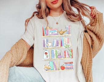 May include: A white t-shirt with a graphic of a bookshelf filled with books, a rose, a lamp, and other whimsical items. The books have titles like "Beauty and the Beast" and "Aladdin".