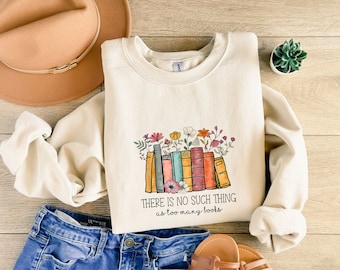Puede incluir: Una sudadera de color crema con un gráfico de una pila de libros coloridos con flores en la parte superior. El texto "THERE IS NO SUCH THING as too many books" está impreso debajo del gráfico.