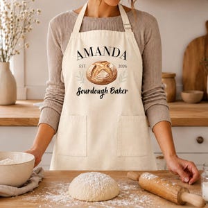 May include: A cream-colored apron featuring a graphic of a round loaf of bread, two sprigs of wheat, and the black text AMANDA, EST. 2026, and Sourdough Baker. The apron includes two front pockets and an adjustable neck strap.