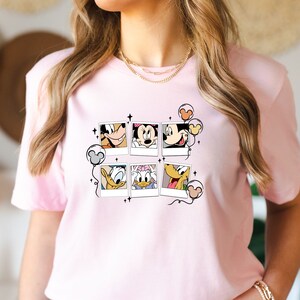 May include: Pink t-shirt with a graphic of Mickey Mouse, Minnie Mouse, Donald Duck, and Goofy in Polaroid frames with balloons.