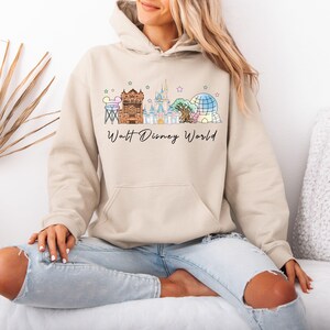 May include: Beige hoodie with a colorful Walt Disney World graphic. The graphic features iconic park landmarks and the text "Walt Disney World" in a script font. The hoodie has a drawstring hood and a front pocket.