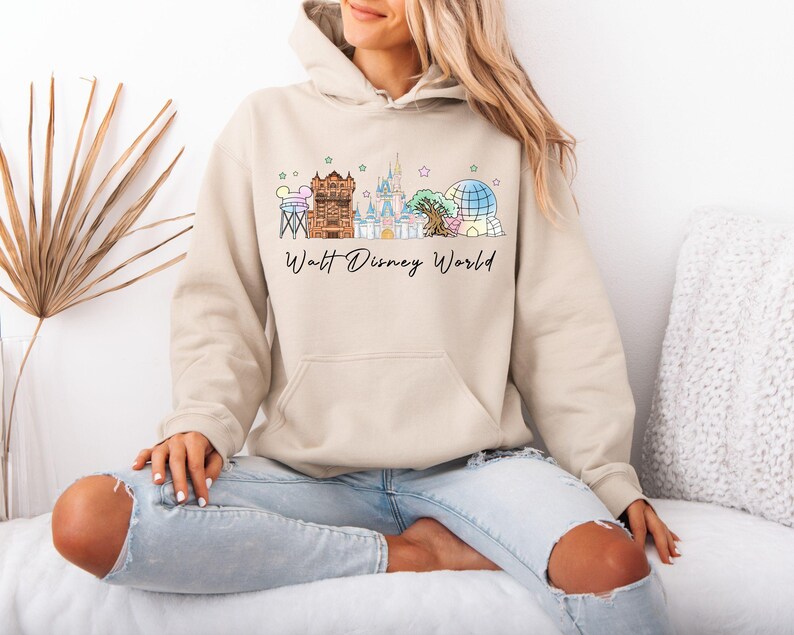 May include: Beige hoodie with a colorful Walt Disney World graphic. The graphic features iconic park landmarks and the text "Walt Disney World" in a script font. The hoodie has a drawstring hood and a front pocket.