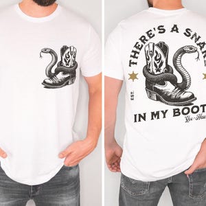 Puede incluir: Camiseta blanca con un dise&ntilde;o en blanco y negro de una bota de vaquero y una serpiente. La parte delantera de la camiseta muestra la bota y la serpiente, y la parte trasera tiene el texto "THERE'S A SNAKE IN MY BOOT".