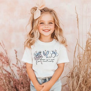 Puede incluir: Una ni&ntilde;a sonriente lleva una camiseta blanca con un dise&ntilde;o caprichoso. La camiseta presenta ilustraciones azules de un personaje, un p&aacute;jaro y un lazo, con el texto "A dream is a wish your heart makes."