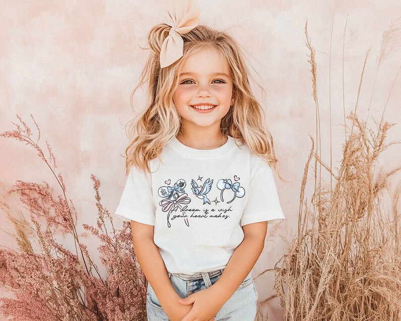 Puede incluir: Una ni&ntilde;a sonriente lleva una camiseta blanca con un dise&ntilde;o caprichoso. La camiseta presenta ilustraciones azules de un personaje, un p&aacute;jaro y un lazo, con el texto "A dream is a wish your heart makes."