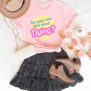 May include: Pink t-shirt with the text "You guys ever think about Dying?" in a retro font on a yellow circle. The shirt is paired with a black skirt with white polka dots and brown ankle boots.