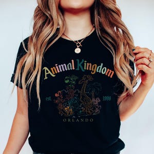 May include: Black t-shirt featuring the words "Animal Kingdom" in rainbow colors, with a graphic of animals and palm trees. The shirt also includes the text "est. 1998 Orlando".