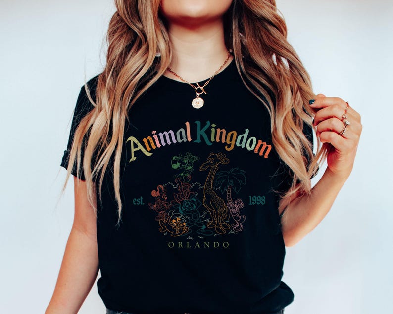 May include: Black t-shirt featuring the words "Animal Kingdom" in rainbow colors, with a graphic of animals and palm trees. The shirt also includes the text "est. 1998 Orlando".