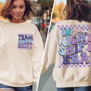 Puede incluir: Una sudadera de color crema con un gráfico morado, azul y blanco que dice "Team Night Shift" en la parte delantera y "In My Night Shift Era" en la parte trasera.