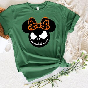 Puede incluir: Camiseta verde con un gr&aacute;fico en blanco y negro de la cara de un personaje y un lazo. El lazo es negro con lunares naranjas. La camiseta est&aacute; anudada a la cintura.