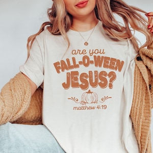 Puede incluir: Camiseta color crema con la frase "are you FALL-O-WEEN JESUS?" en letras marrones. Un diseño de calabaza y la cita bíblica Mateo 4:19 están debajo. La camiseta forma parte de una colección temática de otoño o Halloween.