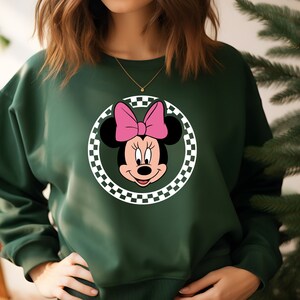 May include: A dark green sweatshirt with a white and black checkered circle featuring a cartoon image of Minnie Mouse with a pink bow.