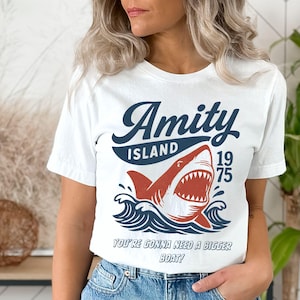 May include: White t-shirt featuring a shark graphic with the words "Amity Island 1975" in navy blue and red. Below the shark, the text reads "You're gonna need a bigger boat!"