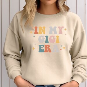 Puede incluir: Una sudadera beige con el texto "In My Gigi Era" en letras retro de colores.