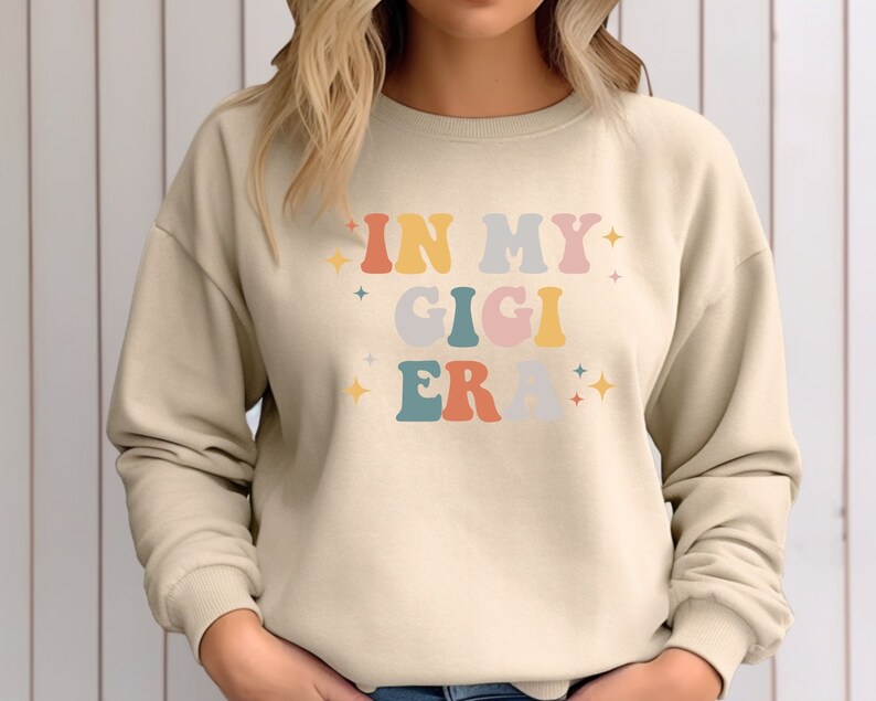 Puede incluir: Una sudadera beige con el texto "In My Gigi Era" en letras retro de colores.