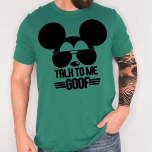 May include: A green short-sleeved t-shirt featuring a black graphic of a mouse silhouette wearing sunglasses. Below the graphic, the text reads "TALK TO ME GOOF" in bold black capital letters, flanked by two horizontal lines on each side of the word GOOF.