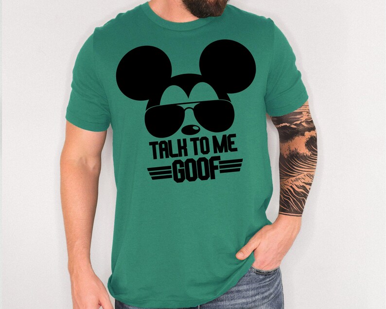 May include: A green short-sleeved t-shirt featuring a black graphic of a mouse silhouette wearing sunglasses. Below the graphic, the text reads "TALK TO ME GOOF" in bold black capital letters, flanked by two horizontal lines on each side of the word GOOF.