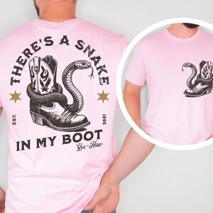 Puede incluir: Camiseta rosa claro con un dise&ntilde;o en blanco y negro de una bota de vaquero y una serpiente. El texto "THERE'S A SNAKE IN MY BOOT" est&aacute; impreso encima y debajo del dise&ntilde;o, con "Yee-Haw" debajo.