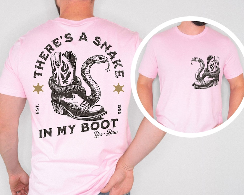 Puede incluir: Camiseta rosa claro con un dise&ntilde;o en blanco y negro de una bota de vaquero y una serpiente. El texto "THERE'S A SNAKE IN MY BOOT" est&aacute; impreso encima y debajo del dise&ntilde;o, con "Yee-Haw" debajo.