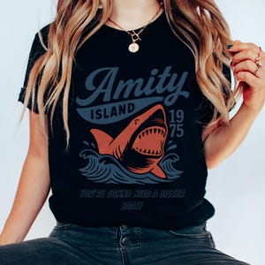 以下が含まれることがあります： 黒いTシャツ。サメのグラフィックデザインで、「Amity Island 1975」と「You're Gonna Need a Bigger Boat！」の文字が入っています。サメはオレンジ色で白い歯があり、青い波が描かれています。