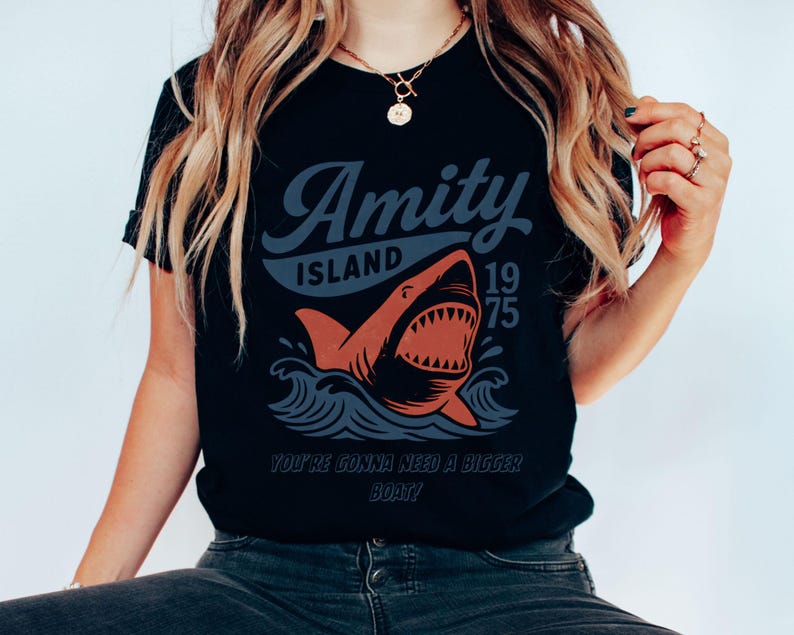 以下が含まれることがあります： 黒いTシャツ。サメのグラフィックデザインで、「Amity Island 1975」と「You're Gonna Need a Bigger Boat！」の文字が入っています。サメはオレンジ色で白い歯があり、青い波が描かれています。