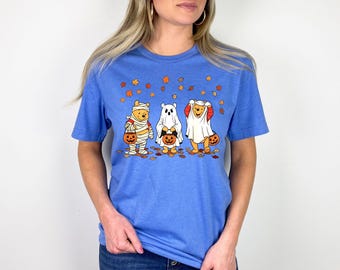 May include: A blue t-shirt with a Halloween design. The graphic shows three cartoon characters in Halloween costumes: a mummy, a ghost, and a character in a sheet, each with a pumpkin. There are autumn leaves scattered around the characters.