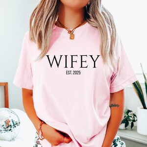 May include: A white ceramic mug with a black handle and the text "But First, Coffee" in a bold, black font. The mug is filled with a dark liquid, likely coffee, and sits on a light-colored surface.