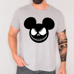 Puede incluir: Una camiseta gris claro con un gr&aacute;fico en blanco y negro. El gr&aacute;fico es una combinaci&oacute;n de orejas de Mickey Mouse y una cara de Jack Skellington. La camiseta est&aacute; hecha de un material suave.
