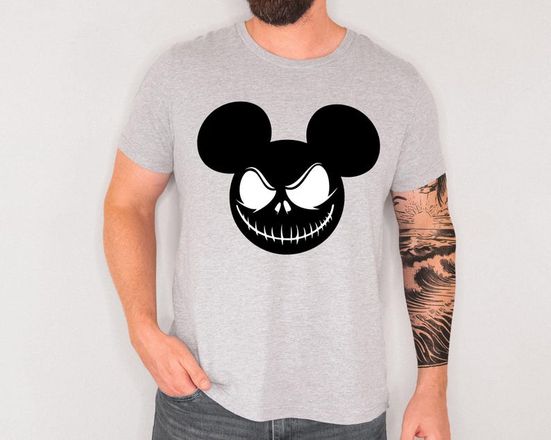 Puede incluir: Una camiseta gris claro con un gr&aacute;fico en blanco y negro. El gr&aacute;fico es una combinaci&oacute;n de orejas de Mickey Mouse y una cara de Jack Skellington. La camiseta est&aacute; hecha de un material suave.