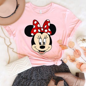 Puede incluir: Camiseta rosa con una imagen de dibujos animados de Minnie Mouse con un lazo rojo con lunares blancos.