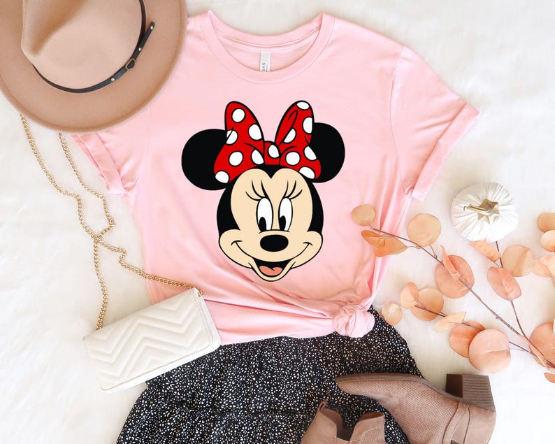 Puede incluir: Camiseta rosa con una imagen de dibujos animados de Minnie Mouse con un lazo rojo con lunares blancos.