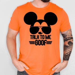 May include: A bright orange short-sleeved t-shirt featuring a black graphic of a mouse silhouette wearing sunglasses. Below the graphic, the text reads "TALK TO ME GOOF" in bold black capital letters, flanked by two horizontal black lines.