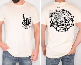 Può includere: T-shirt color crema con disegni grafici neri. La parte anteriore presenta una mano di scheletro che fa un gesto rock and roll con la parola "FATHERHOOD". Il retro mostra uno scheletro che beve birra con il testo "Surviving Fatherhood One Beer at a Time".