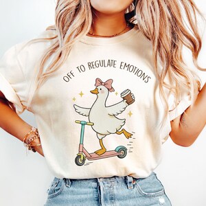 Puede incluir: Una camiseta color crema con un gráfico de un ganso blanco con un lazo rosa, montado en un patinete rosa y azul mientras sostiene una taza de café. El texto sobre el ganso dice OFF TO REGULATE EMOTIONS en una fuente negra arqueada.
