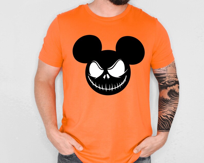 Puede incluir: Camiseta naranja brillante con un gr&aacute;fico en blanco y negro de un personaje inspirado en Mickey Mouse con una sonrisa siniestra. El dise&ntilde;o combina elementos de Mickey Mouse con un tema de Halloween. La camiseta es de manga corta.