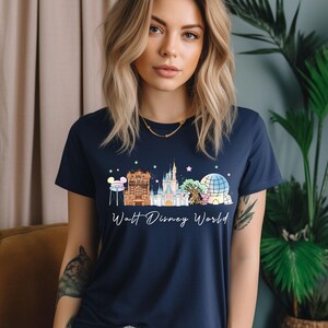 May include: Navy blue t-shirt featuring a colorful illustration of Walt Disney World landmarks. The design includes Cinderella's castle, Spaceship Earth, and other iconic structures. The words "Walt Disney World" are written in a script font below the illustration.