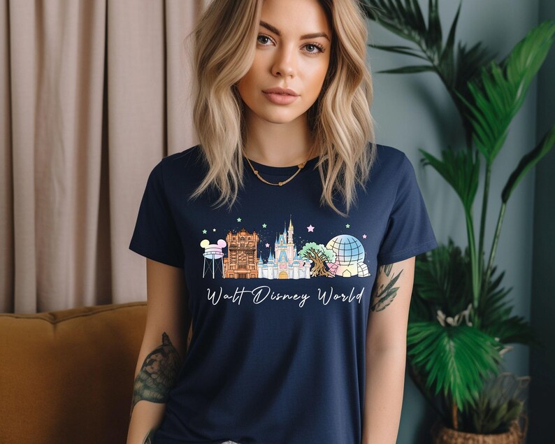 May include: Navy blue t-shirt featuring a colorful illustration of Walt Disney World landmarks. The design includes Cinderella's castle, Spaceship Earth, and other iconic structures. The words "Walt Disney World" are written in a script font below the illustration.