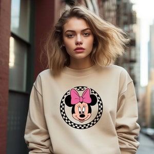 May include: A beige sweatshirt with a black and white checkered circle featuring a cartoon image of Minnie Mouse with a pink bow.