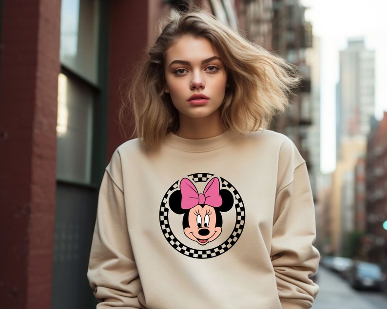 May include: A beige sweatshirt with a black and white checkered circle featuring a cartoon image of Minnie Mouse with a pink bow.