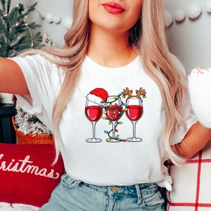 May include: White t-shirt featuring a graphic of three wine glasses filled with red wine. One glass has a Santa hat, another has reindeer antlers, and all are decorated with Christmas lights. The shirt is a festive holiday design.