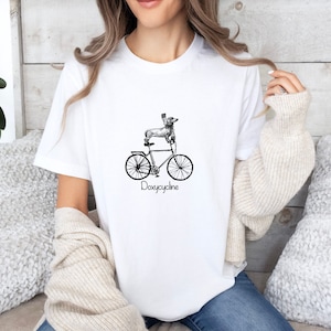 May include: White t-shirt with a black and white illustration of a dachshund riding a bicycle. The text "Doxycycline" is printed below the image.