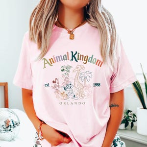 May include: A light pink t-shirt featuring the text "Animal Kingdom" in a multi-colored font, with a graphic of various cartoon animals. Below the text are the words "est. 1998" and "Orlando".